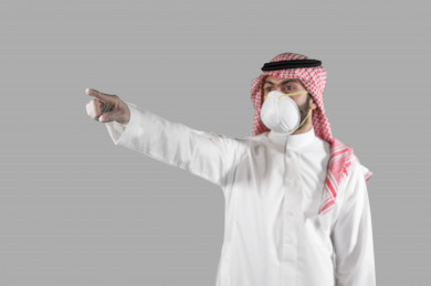 بورتريه لشاب سعودي يرتدي الكمامة و القفازات ، يرفع يده و يؤشر باصبعة على مكان ، تجنب انتشار العدوى ، الحفاظ على المسافات ، خلفية بيضاء
