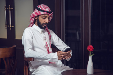 رجل عربي سعودي ، يمسك بيده هاتفه المحمول ، وبجانبه  فنجان القهوة  ، طلب القهوه في متجر القهوه