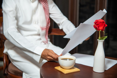 رجل أعمال عربي سعودي سعيد ،يعمل في المقهى و يحمل بيده أوراق رسمية ، بجانبه  فنجان القهوة ،  متجر القهوه