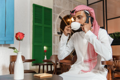 رجل عربي سعودي ، يستقبل مكالمة هاتفية في المقهى ، يحمل بيده فنجان القهوة  ، طلب القهوه في متجر القهوه