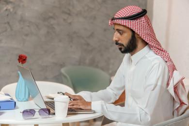رجل أعمال عربي سعودي سعيد ، يعمل على الكمبيوتر المحمول ، بجانبه  فنجان القهوة ،  متجر القهوه